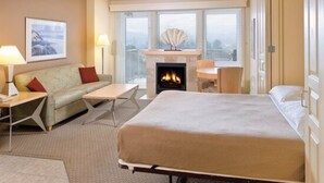Ett hotellrum med en säng, en soffa, ett litet bord, en öppen spis och utsikt över staden genom stora fönster.