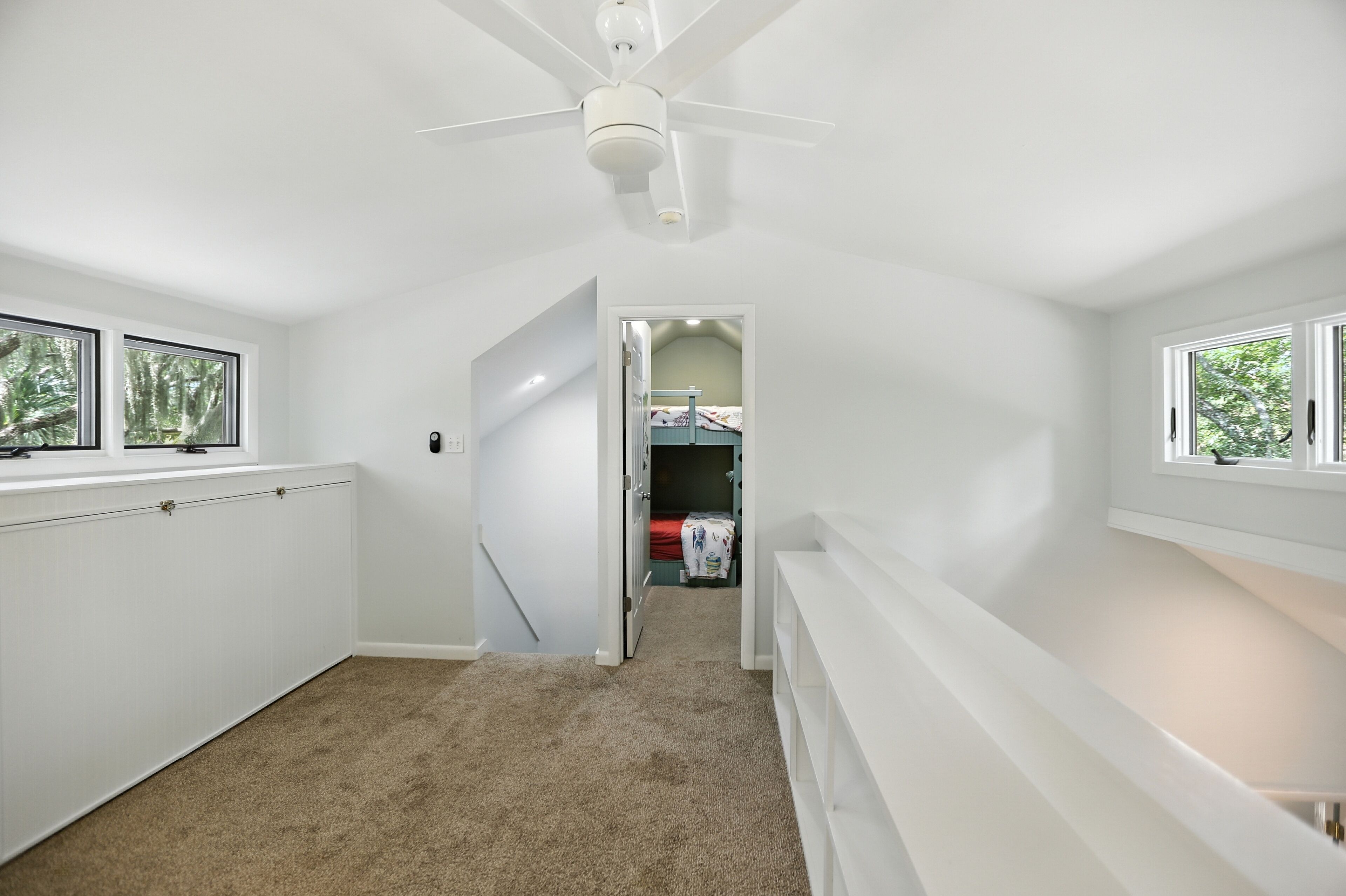 A narrow hallway with a ceiling fan, white walls, and carpeted floor leading to a bedroom with a bed and a window.