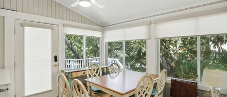A dining area with a wooden table and chairs, large windows with a view of trees, and a ceiling fan.