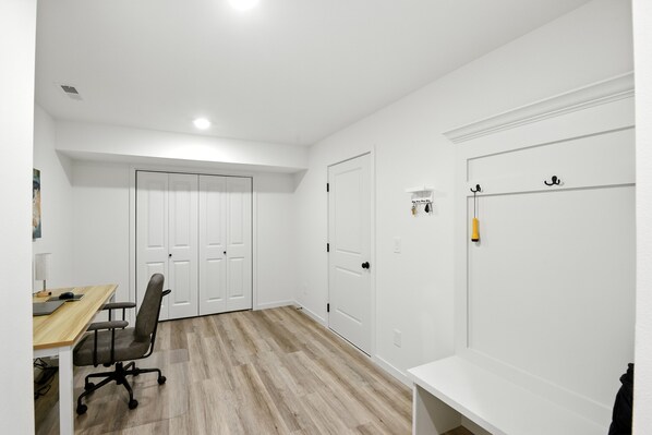 A room with a wooden floor, a desk with a chair, and white walls. There are two white doors and a coat rack with hooks.