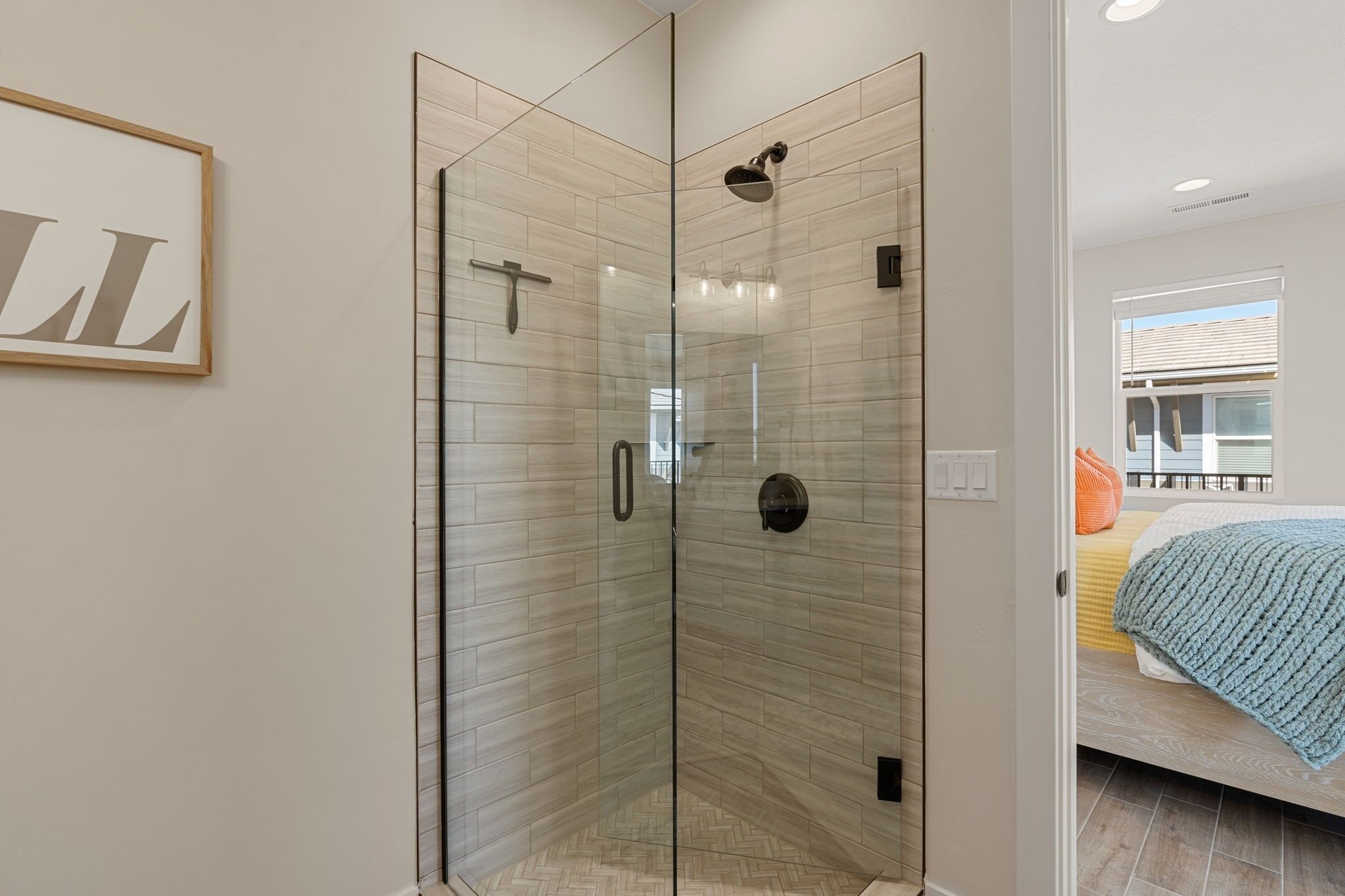 A modern bathroom with a glass shower enclosure, a framed artwork on the wall, and a visible bedroom with a bed and a window.