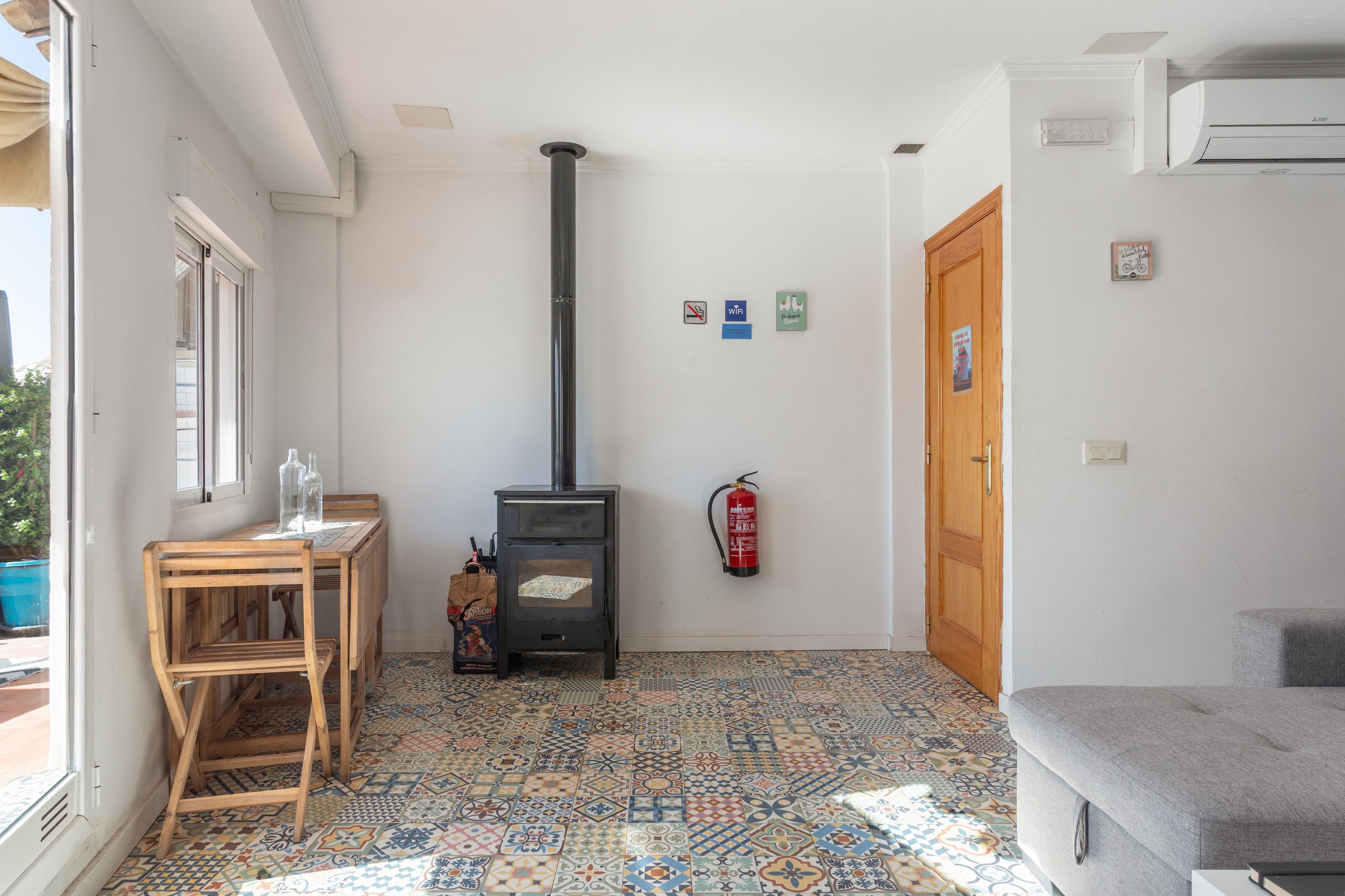 Ein Zimmer mit Fliesenboden, einem Holztisch mit Stühlen, einem schwarzen Herd, einem Feuerlöscher und einer Tür mit Holzrahmen.