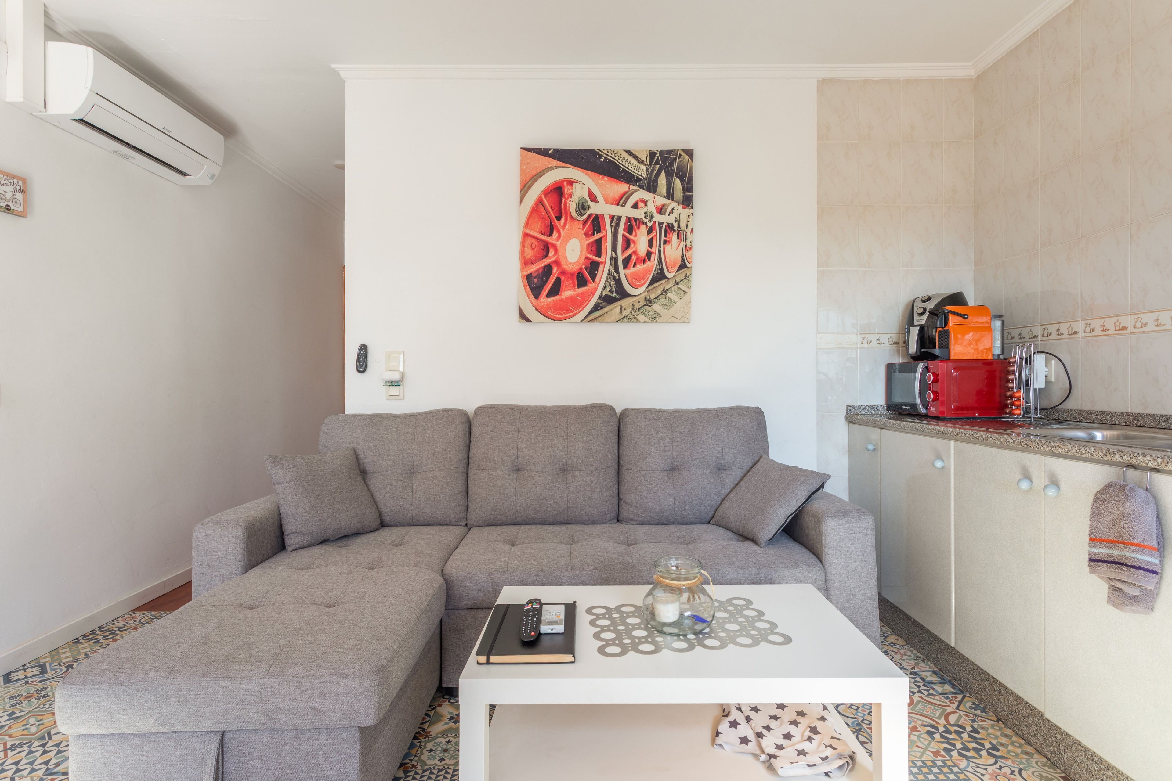 Un espace de vie moderne comprenant un canapé d’angle gris, une table basse blanche et une cuisine équipée d’un four à micro-ondes et d’une bouilloire.