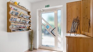 Un espace d’accueil avec une étagère en bois où sont exposées des brochures, une porte en verre ornée d’un motif géométrique et un petit vase contenant des plantes séchées.