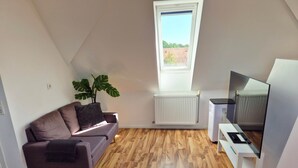 En moderne stue med trægulv, en grå sofa, en stor plante, en hvid radiator og et fladskærms-tv på et hvidt stativ.