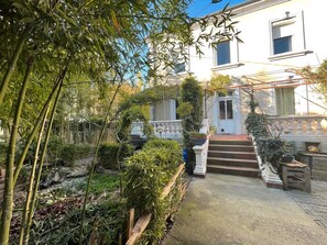 A garden with bamboo, a white building with a staircase, and a small wooden bench. - Bed and Breakfast 'Chambre Spacieuse' with Shared Terrace, Shared Garden and Wi-Fi (Le Teil)