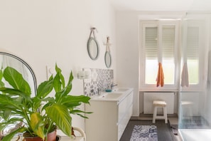 A bathroom with a white sink, two mirrors, a potted plant, a radiator, and a window with blinds.