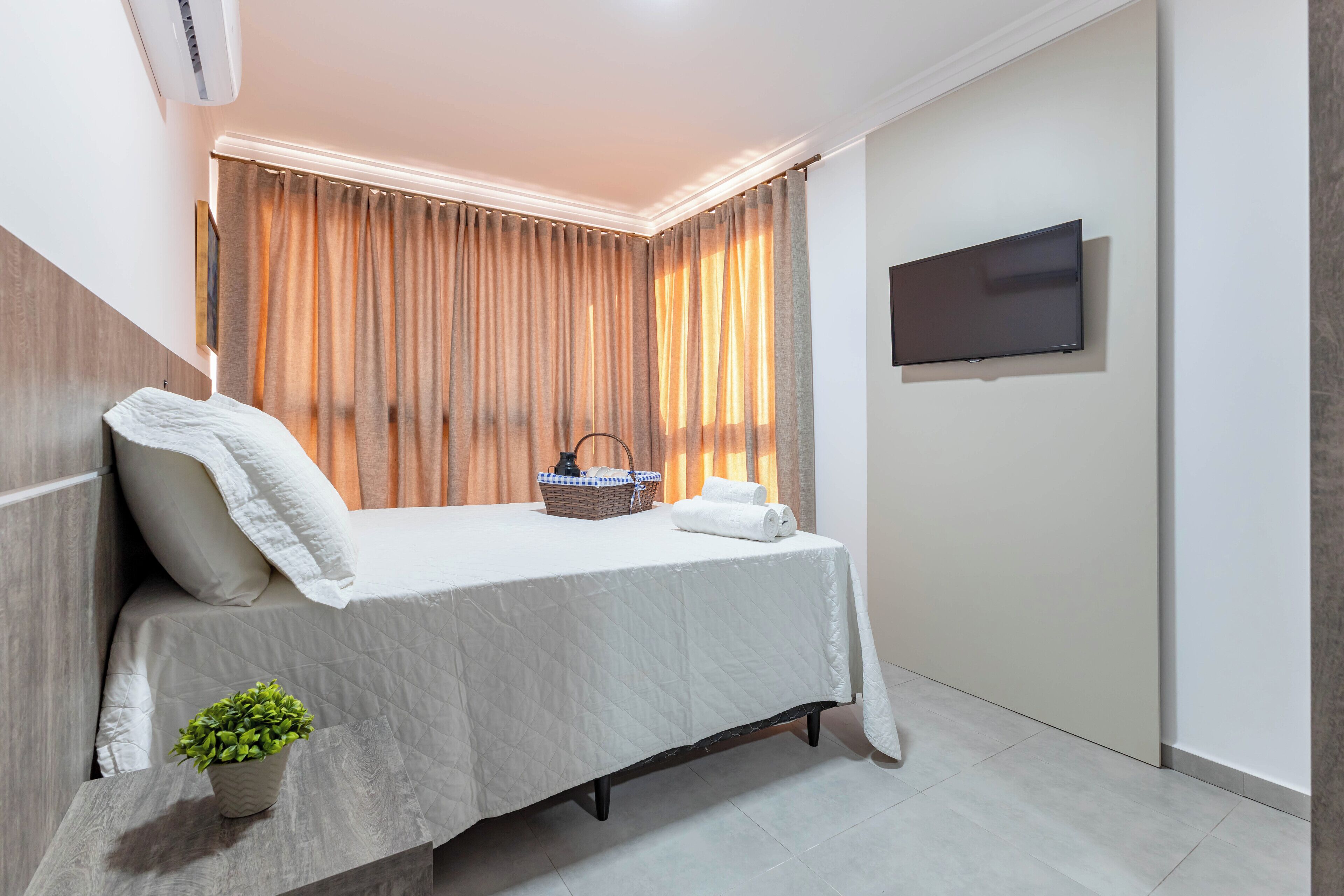 Un cuarto de hotel ordenado con una cama, un televisor de pantalla plana, una planta pequeña y una cesta en la mesita de noche.