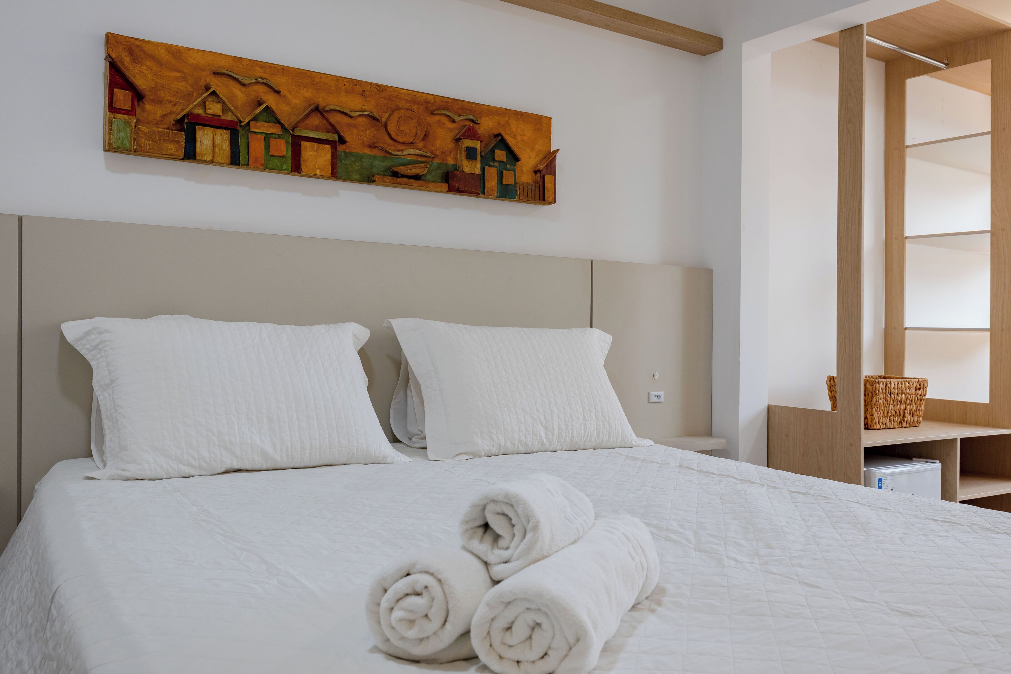 A neatly made bed with white linens and rolled towels, a wooden headboard, and a painting of houses on the wall.