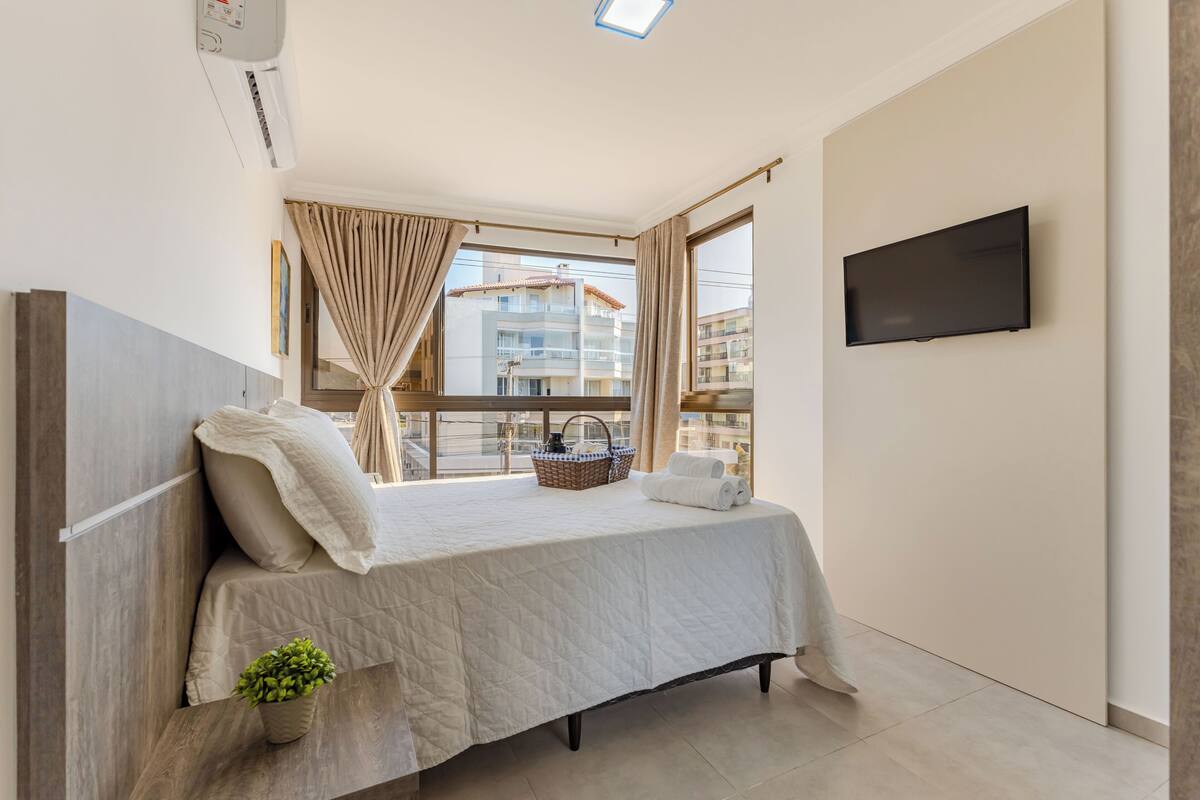 Une chambre avec un lit, une télévision, une fenêtre avec des rideaux, une vue sur le balcon et une petite plante sur la table de chevet.