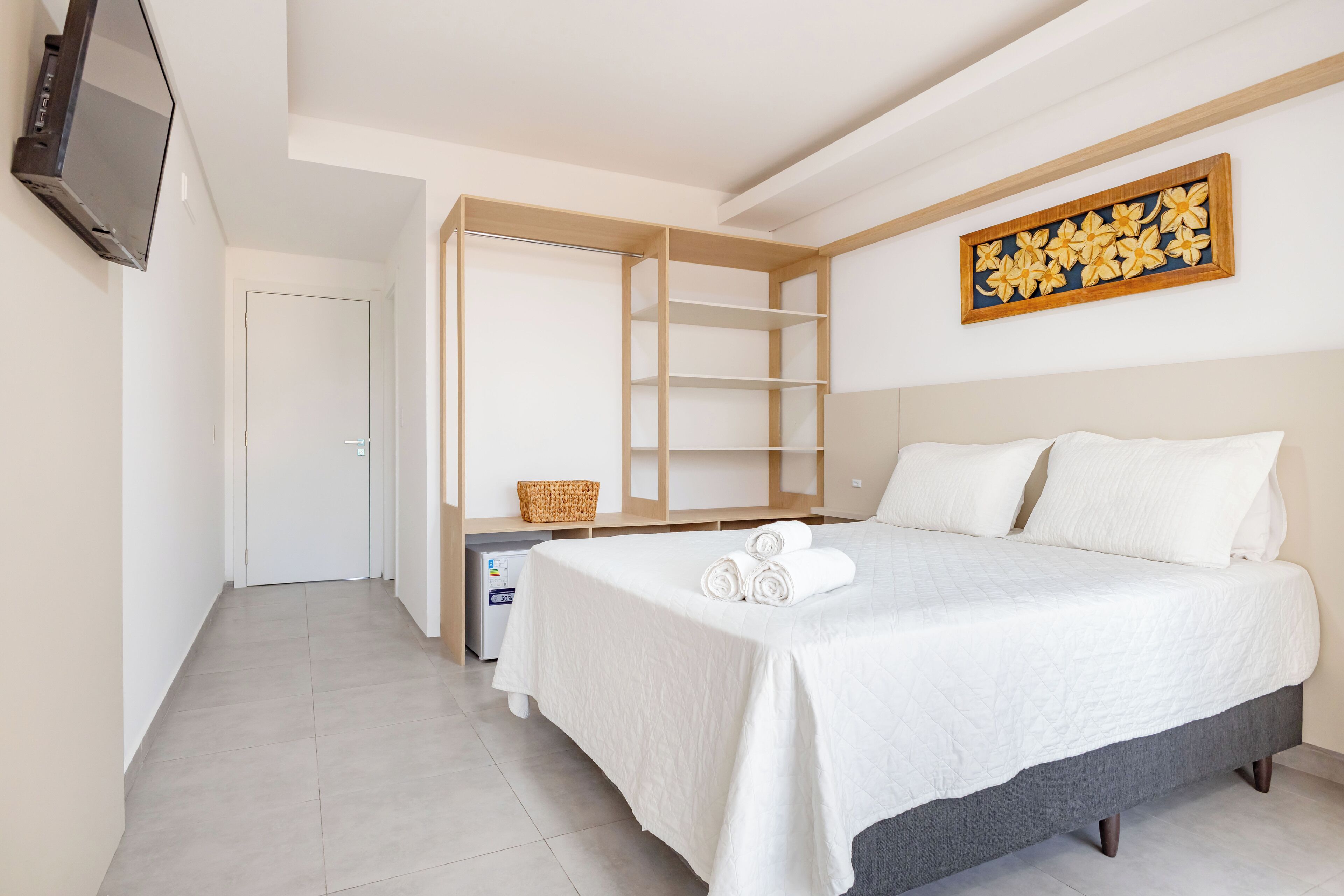 Habitación de hotel con cama, televisión montada en la pared, escritorio con cesta y armario con estantes.