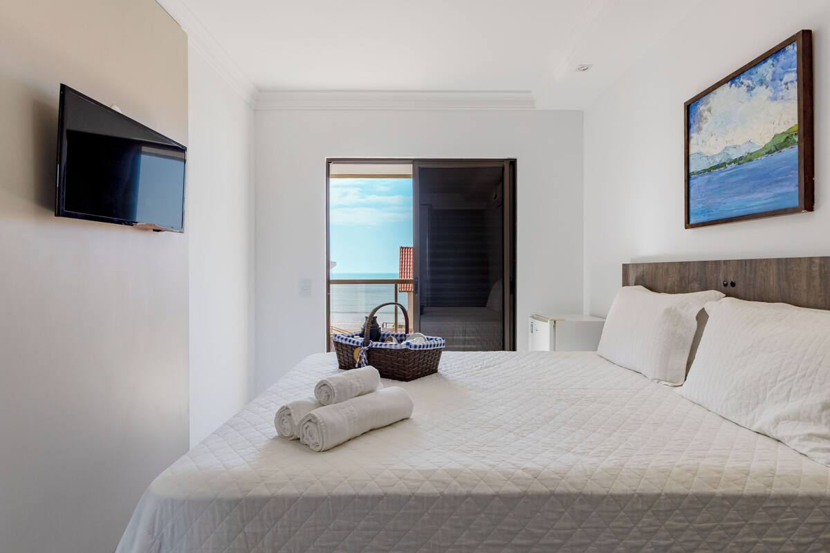 Une chambre d’hôtel avec un lit, un téléviseur à écran plat, un tableau et une vue sur la mer, visible par une porte ouverte.