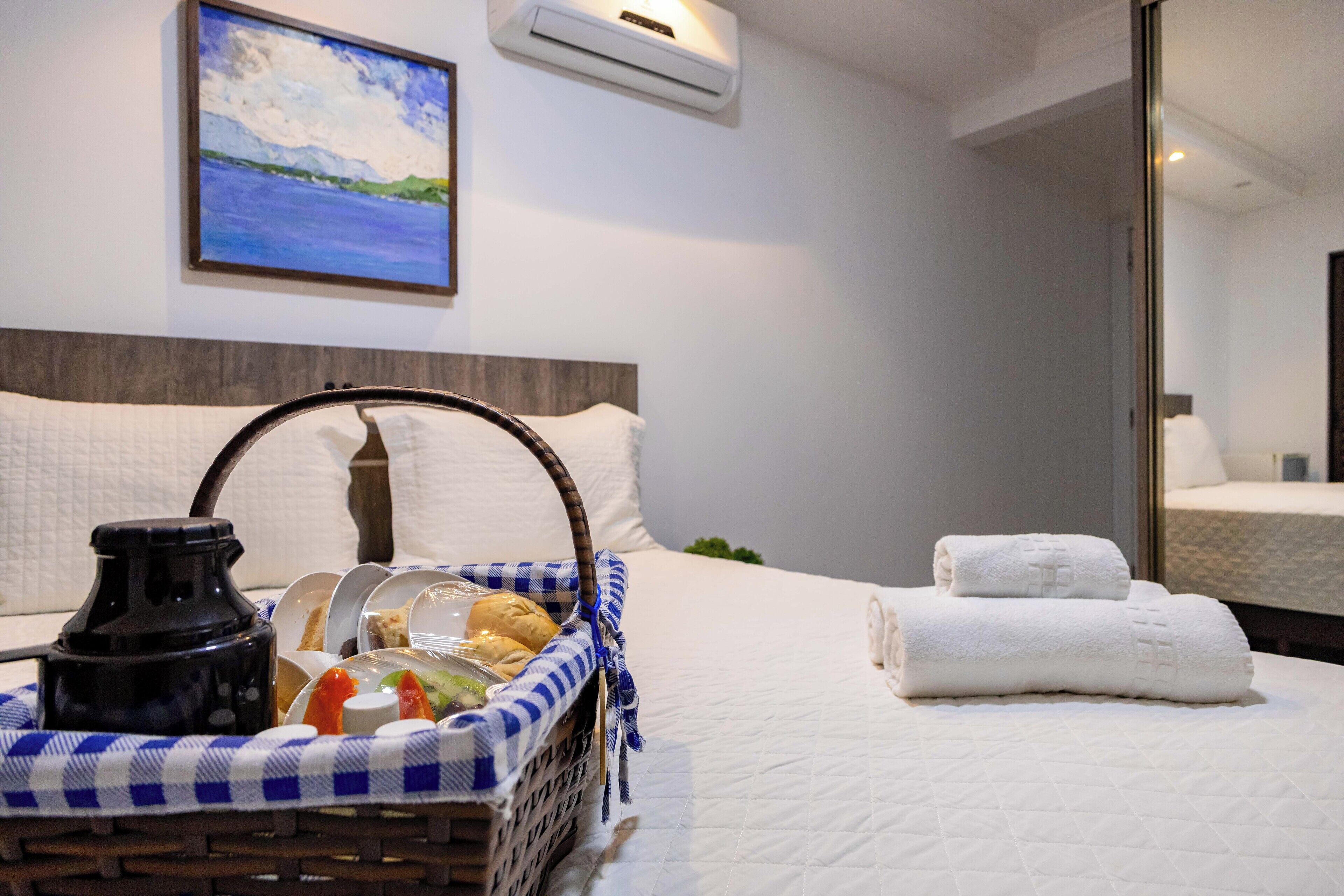 A neatly made bed with white linens, a basket of breakfast items, and a painting of a landscape on the wall.