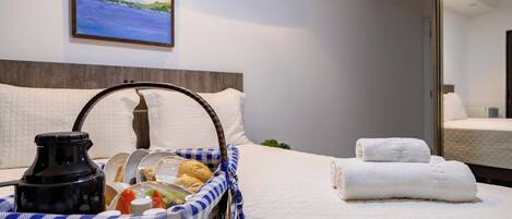 A neatly made bed with white linens, a basket of breakfast items, and a painting of a landscape on the wall.