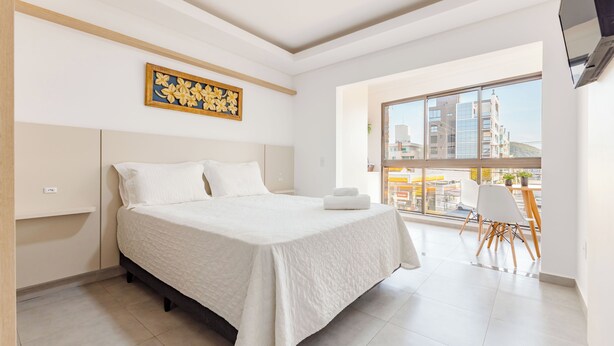 A modern bedroom with a large bed, a view of the city through a large window, and a small dining area with chairs. - Kia Ora Suítes (Bombinhas)