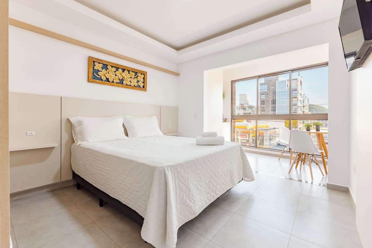 Une chambre moderne avec un grand lit, une vue sur la ville grâce à une grande fenêtre et un petit coin repas avec des chaises.
