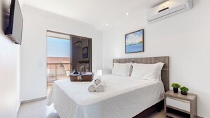 A modern bedroom with a large bed, a nightstand, a TV, and a view of the outside balcony. - Kia Ora Suítes (Bombinhas)