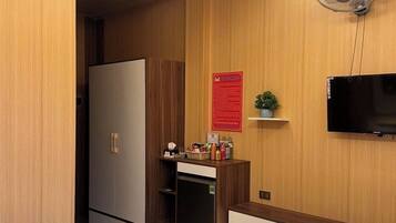 A modern hotel room with a wooden panel wall, a flat-screen TV mounted above a wooden console, a minibar, and a small plant on the console.