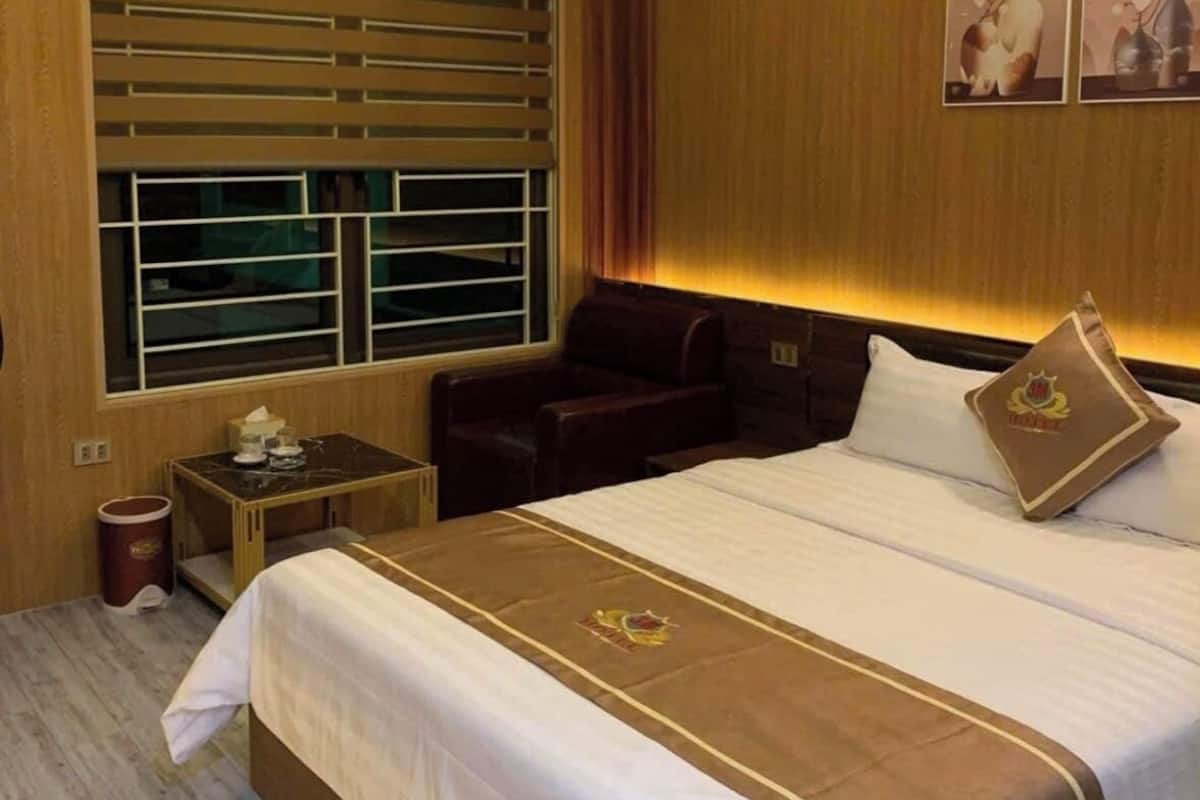 Habitación de hotel con una cama grande, cabecera de madera, una mesita con un florero y una ventana con persianas.