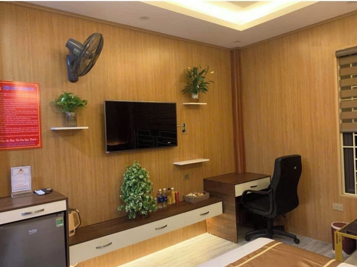 Une chambre d’hôtel avec un mur recouvert de panneaux de bois, un téléviseur à écran plat fixé au-dessus d’un bureau, un fauteuil de bureau noir, une petite plante en pot et un bureau avec des tiroirs.