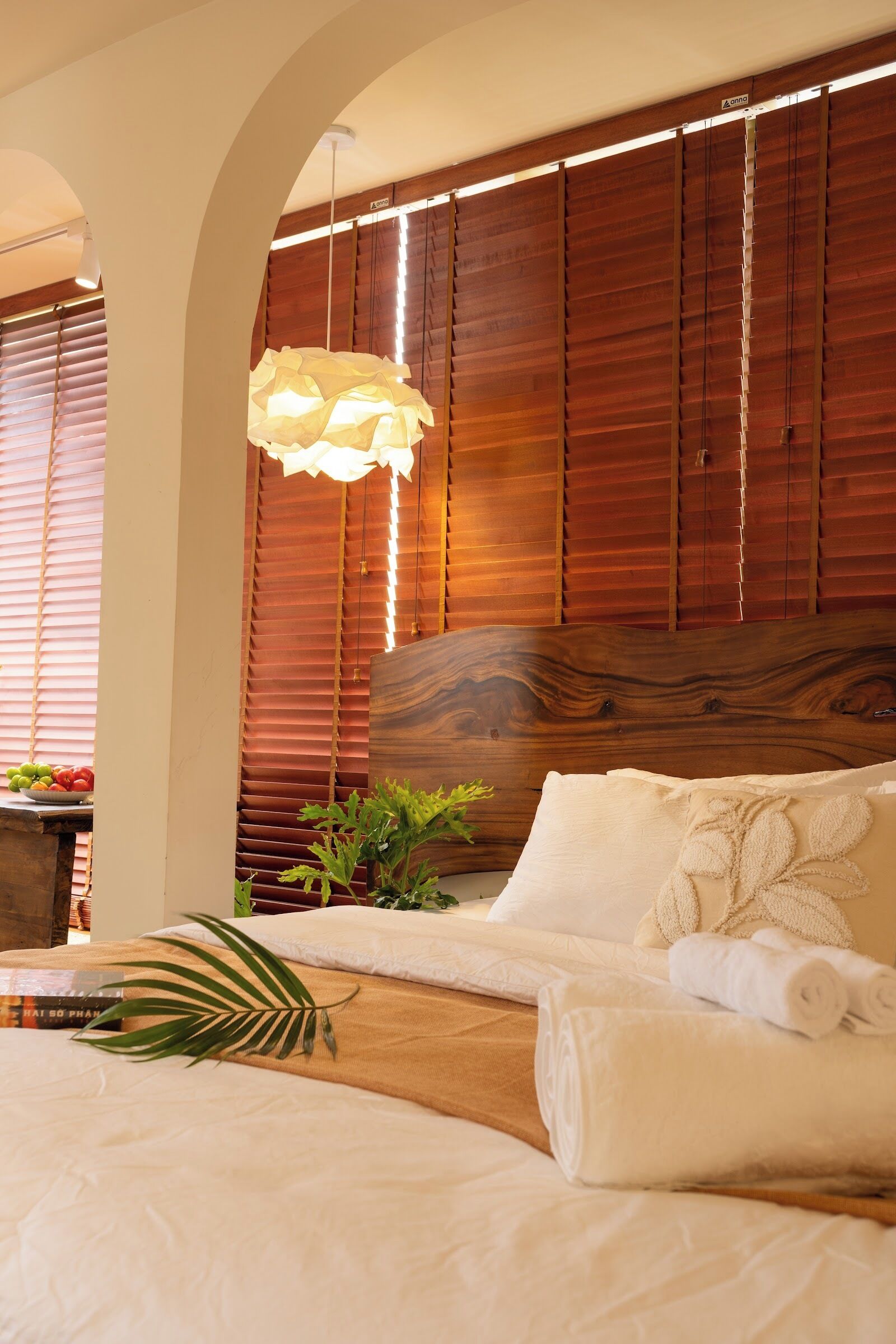 A bedroom with a wooden headboard, a hanging light fixture, a bed with white linens, and a plant on the bedside table.