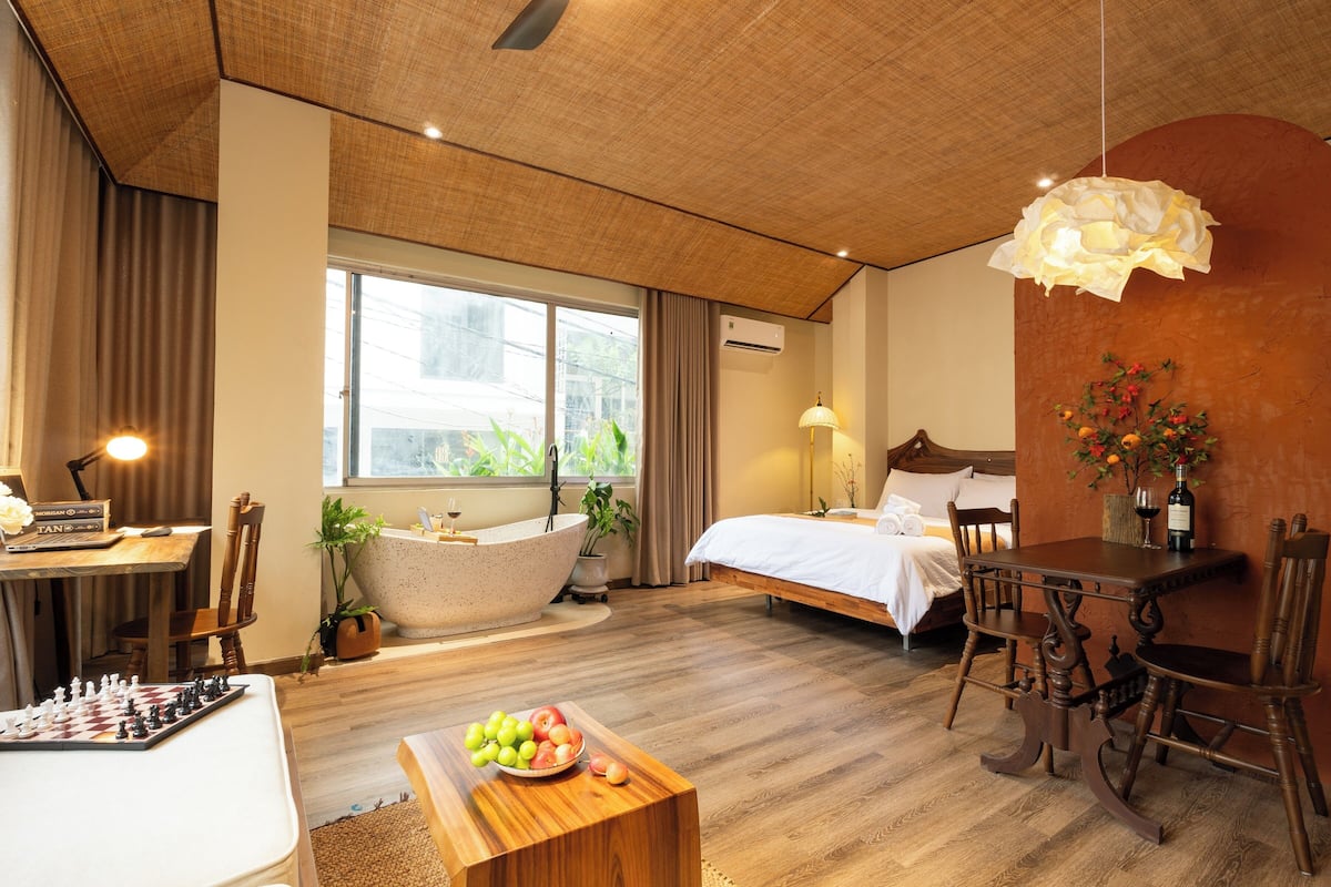 Habitación de hotel con un ventanal grande, una bañera, una mesa de comedor con sillas, un tablero de ajedrez y un frutero.