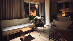Una sala de estar acogedora con un sofá, una mesa de centro de madera y una silla. Hay una ventana con cortinas, un ventilador de techo y una planta.