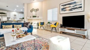 A modern living room with a sofa, coffee table, and TV. There is a fireplace, a dining area, and a kitchen in the background. - Relaxing 4BR Retreat w/ Refreshing Pool (Bermuda Dunes)
