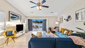 A spacious living room with a blue sofa, a yellow armchair, a flat-screen TV, and a ceiling fan. - Relaxing 4BR Retreat w/ Refreshing Pool (Bermuda Dunes)