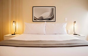 A neatly made bed with white linens, two bedside lamps, and a framed abstract artwork above the bed.