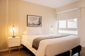 A neatly made bed with a white comforter and a gray throw blanket, a bedside table with a lamp, a framed abstract artwork on the wall, and a window with a white blind.