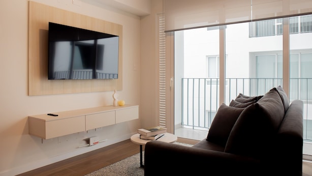 A modern living room with a flat-screen TV mounted on the wall, a sofa, a small side table, and a large window with blinds. - w Charming 2BR Stay in Miraflores (Lima)