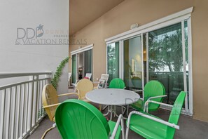 A balcony with a round table and green and yellow chairs, a white railing, and a glass door leading to a room. - Legacy Tower II 106 | Charming Ground-Floor Unit! (Gulfport)