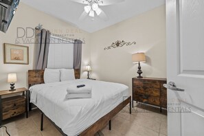 A bedroom with a bed, bedside tables, a lamp, a ceiling fan, and a wall-mounted decoration. - Legacy Tower II 106 | Charming Ground-Floor Unit! (Gulfport)