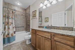 A bathroom with a bathtub, toilet, vanity with sink, wooden cabinets, and a shower curtain with sea creature prints. - Legacy Tower II 106 | Charming Ground-Floor Unit! (Gulfport)