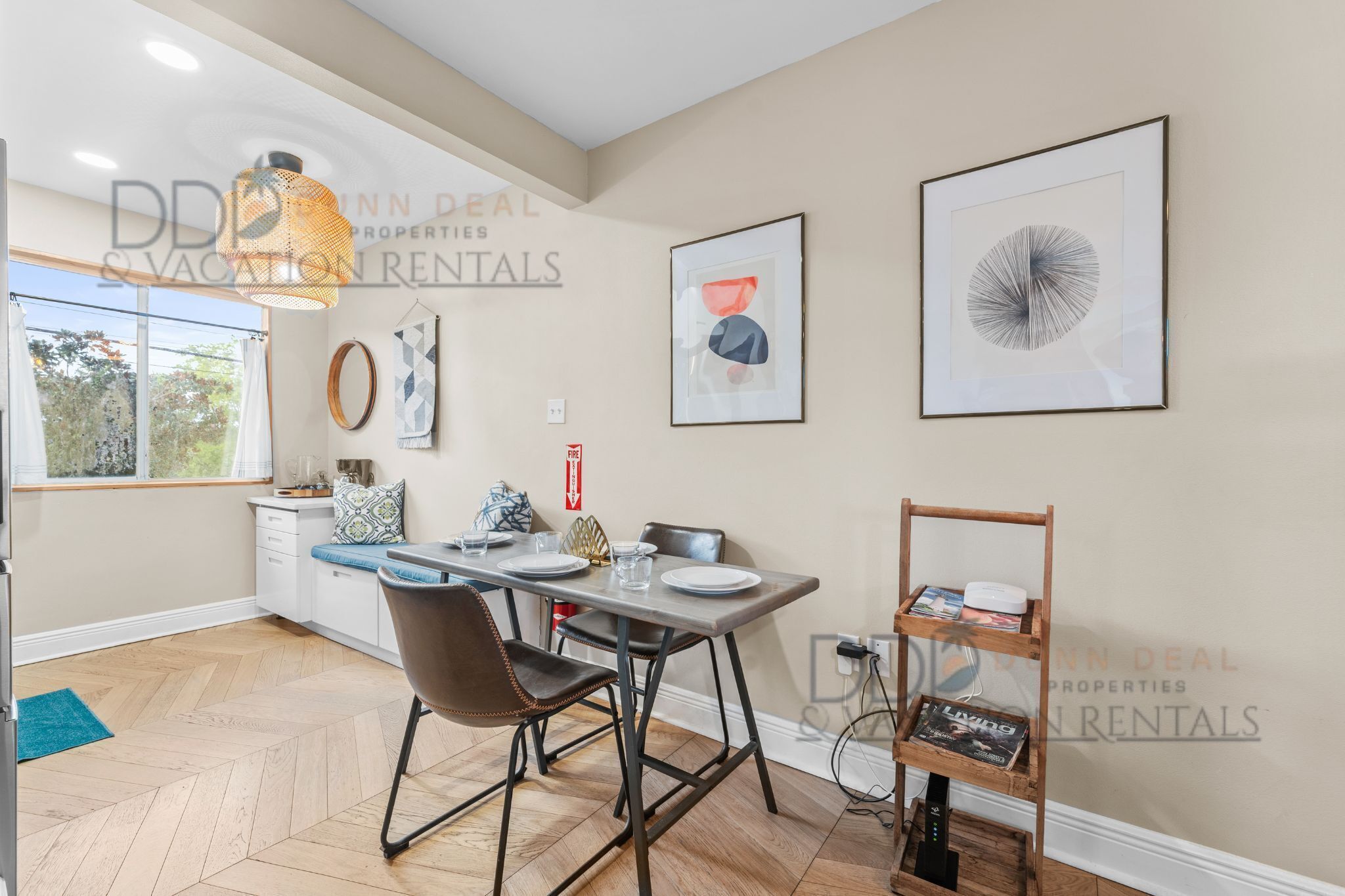 Un coin repas comprenant une table, des chaises et un luminaire suspendu. Des œuvres d’art encadrées ornent le mur, et une petite étagère contient des livres.