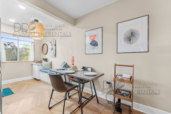 Un coin repas comprenant une table, des chaises et un luminaire suspendu. Des œuvres d’art encadrées ornent le mur, et une petite étagère contient des livres.