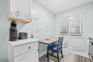 Una kitchenette con cafetera, tostadora y extintor. Hay una ventana con persianas y una silla con cojín azul.