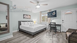 Ein Schlafzimmer mit Bett, Schreibtisch und Stuhl. Es gibt einen Deckenventilator, ein Fenster mit Vorhängen und eine Tür.