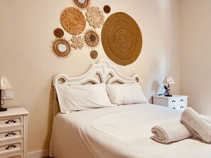 A neatly made bed with white linens, a white nightstand with a lamp, and decorative woven wall art. - Cosmópolis 2 (Santa Lucía de Tirajana)