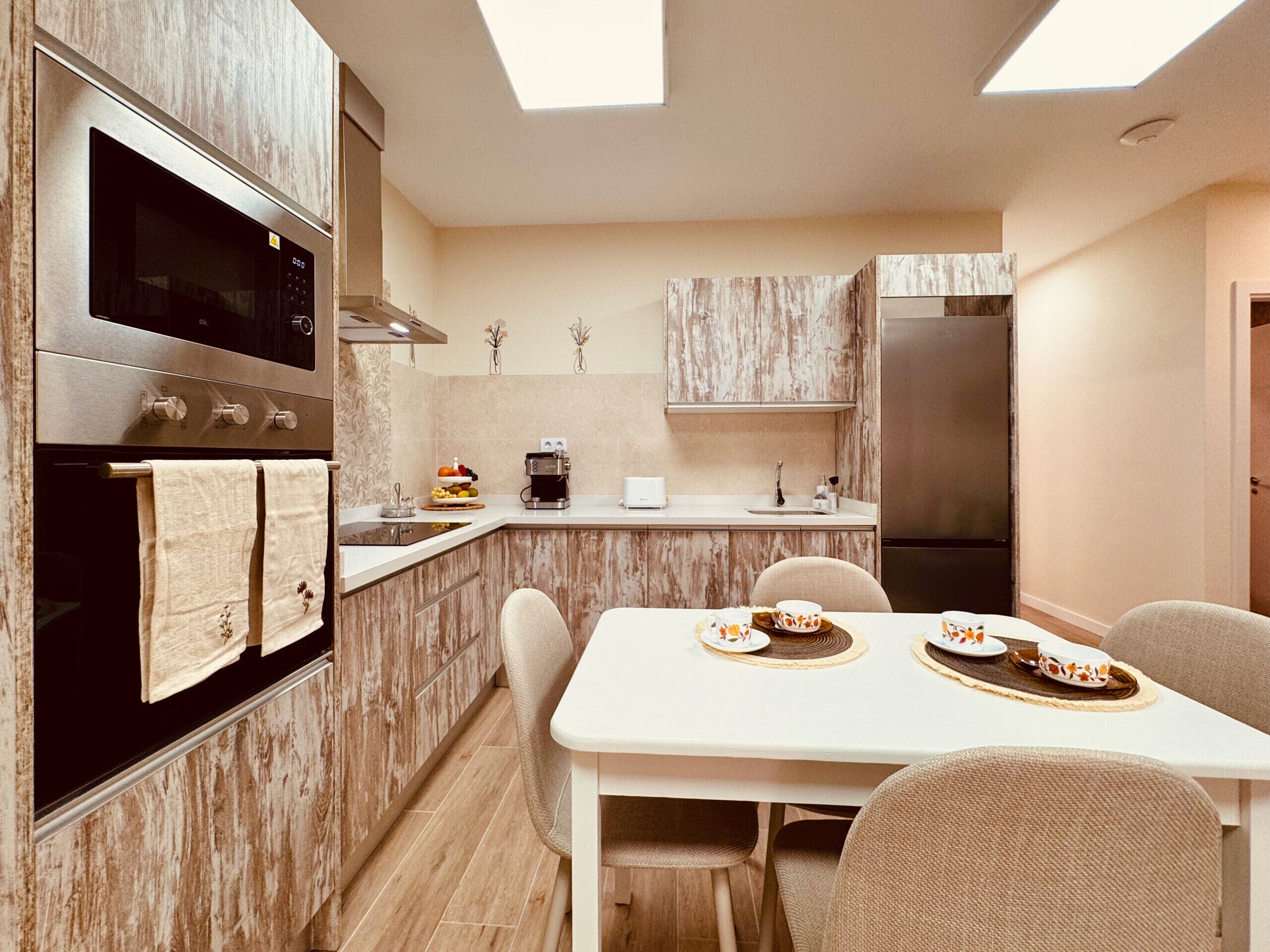 A modern kitchen with a built-in oven, microwave, and stainless steel refrigerator. There is a dining table with chairs and a small island with a coffee machine.