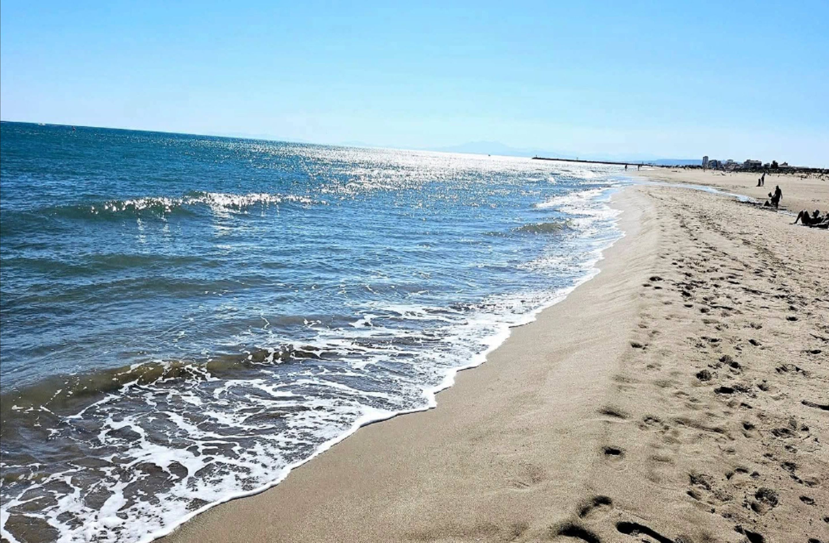 A beach with clear blue water, sandy shore, and visible footprints.