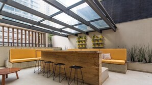 A modern interior with a glass ceiling, a wooden bar counter, yellow seating, and black stools. - Uwin Brooklin NR (São Paulo)
