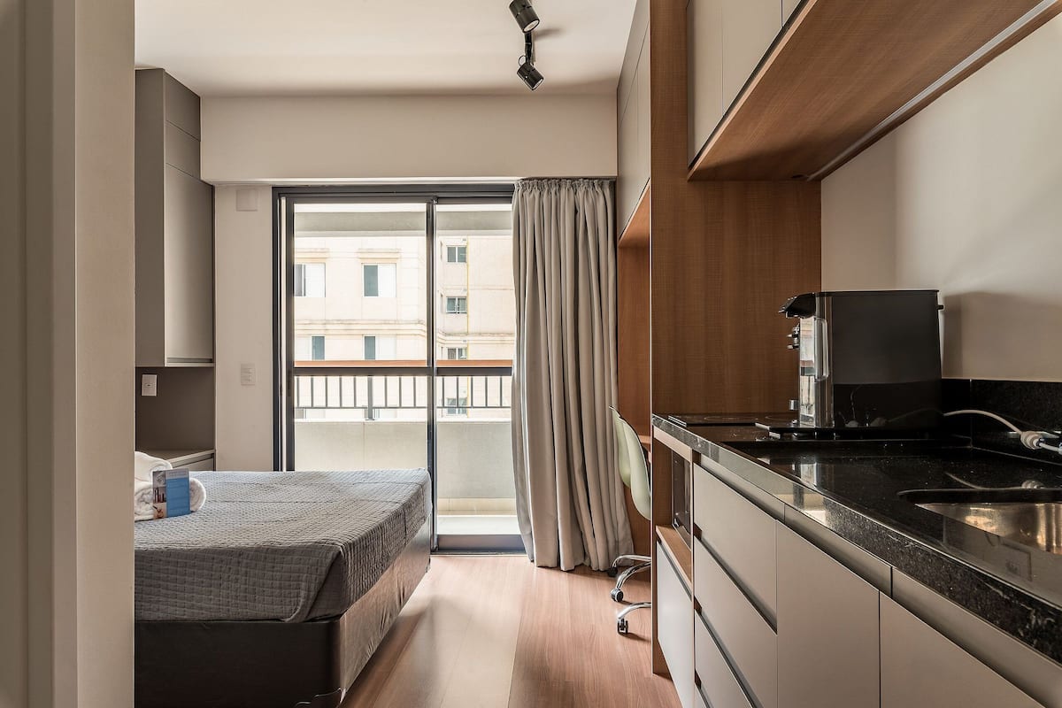 A compact hotel room with a kitchenette, a bed, a sliding glass door to a balcony, and a view of the cityscape.