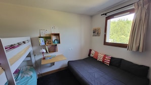 A small room with a bunk bed, a desk with books and a lamp, a sofa, and a window with curtains.