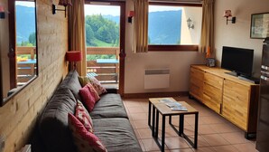 A cozy living room with a sofa, a coffee table, a TV, and a view of mountains through large windows.