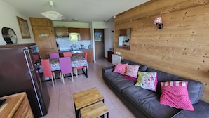 A modern living room with a grey sofa, wooden coffee table, and pink and white patterned cushions. A kitchen with wooden cabinets and a dining area with pink chairs and a table are visible.