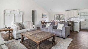 Un vaste salon comprenant une grande table basse en bois, deux canapés gris, un canapé blanc et un coin cuisine avec des meubles blancs et une fenêtre.