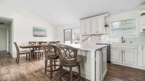 Une cuisine avec des meubles blancs, un coin repas avec des chaises en bois et une grande fenêtre.