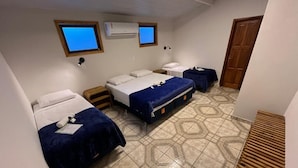 Una habitación pequeña y funcional con tres camas, cada una con cubrecama azul y almohadas blancas. Cuenta con dos unidades de aire acondicionado fijadas a la pared, un banco de madera y piso de baldosas con diseño geométrico.